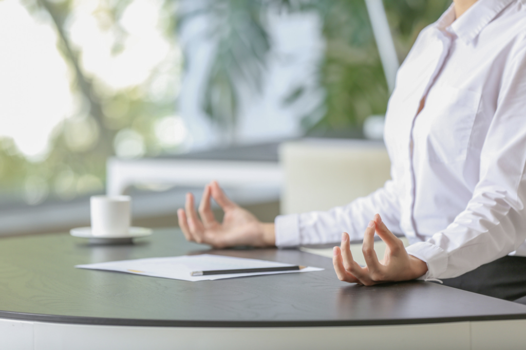 A business woman sits at her desk, hands poised in a meditation mudra next to her coffee cup.